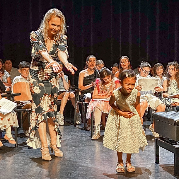 A woman in a floral dress gesturing toward a young girl in a white dress on stage